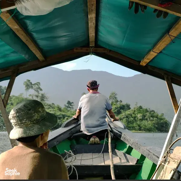 On-deck view from a small boat on a river through Kuching’s rainforest, with travellers watching forested hills