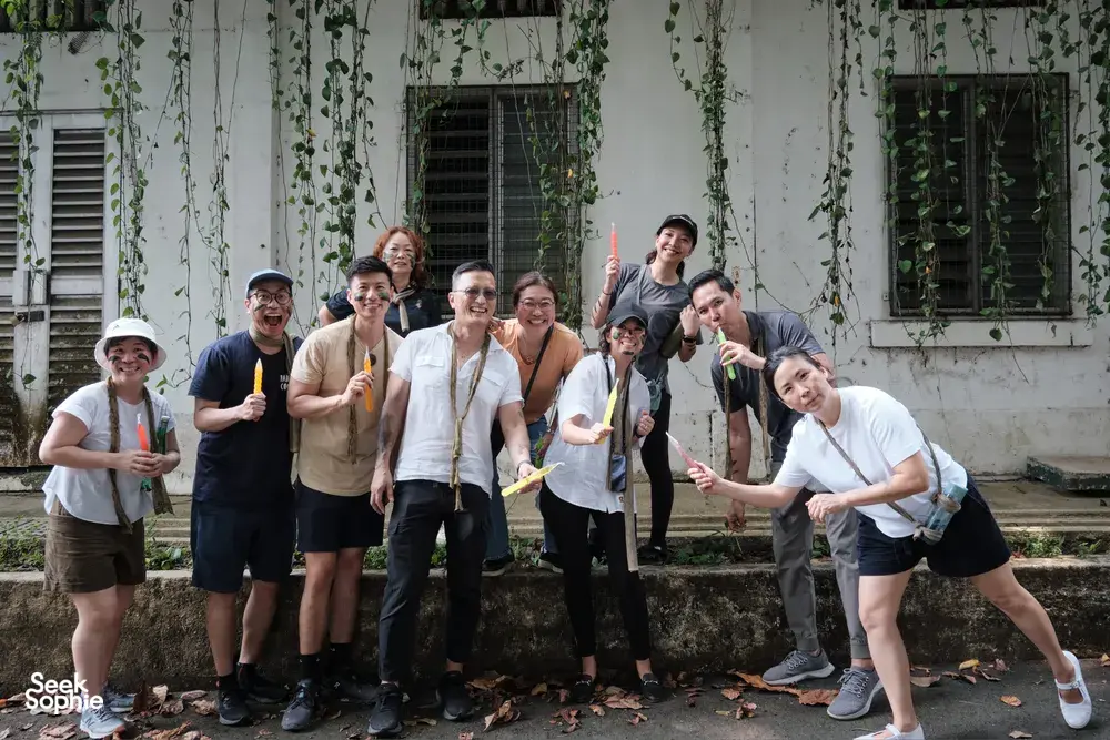 Team adventure group posing outdoors at Fort Serapong in Singapore, smiling with drinks in hand in front of a vine-covered wall