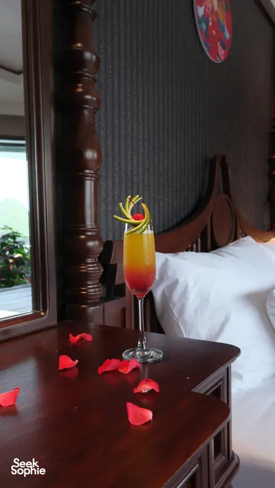 Cat Ba cruise cabin turndown with rose petals on the bedside table and a tropical cocktail drink beside the made bed