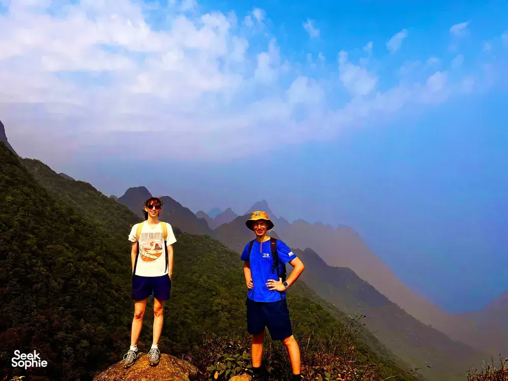 Two hikers pose on a ridge with layered mountain silhouettes behind them on Mt Fansipan in Sapa during a 1-day eco trek