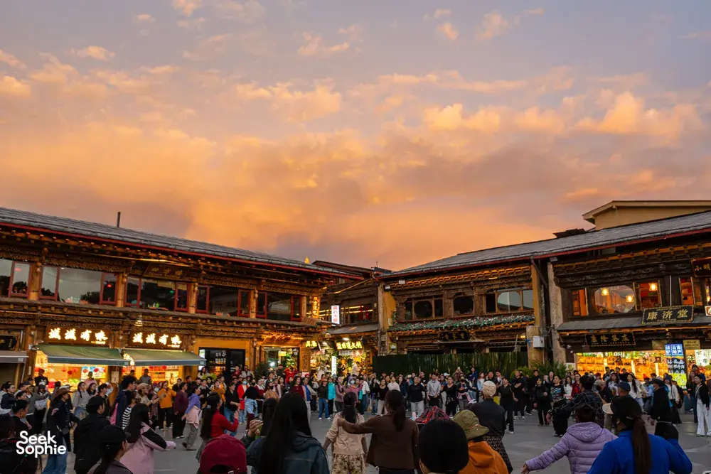 Crowd gathers in Shangri-La’s old town at dusk, with illuminated traditional buildings lining a central square beneath orange sky