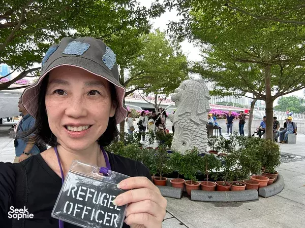 Participant from the Survive The Collapse sustainability outdoor scavenger hunt in Singapore, smiling while holding a “REFUGEE OFFICER” card
