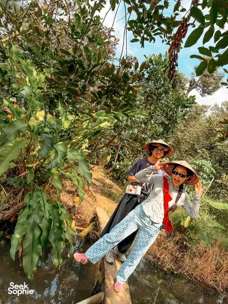 Tour guests in conical hats standing on a muddy Mekong Delta track beside fruit trees, smiling while exploring the plantation
