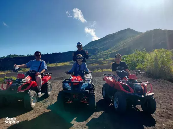 Helmeted ATV riders pause on a black-sand track as the volcano looms during a Bali adventure, at Mt Batur