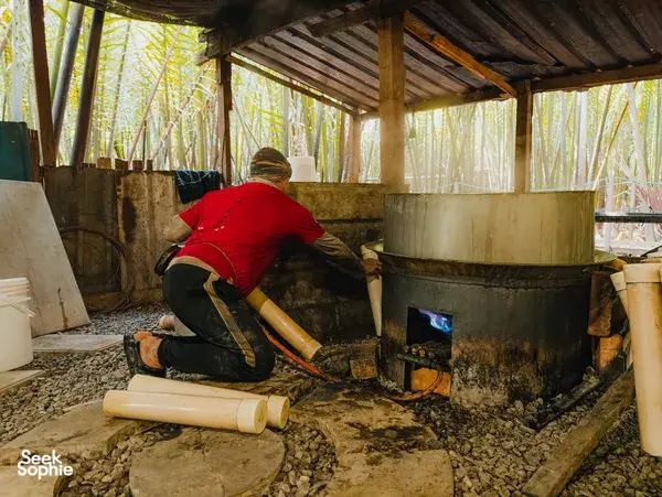 Worker crouched by a large cauldron, boiling palm sap under a bamboo shelter in Kuching, Sarawak workshop