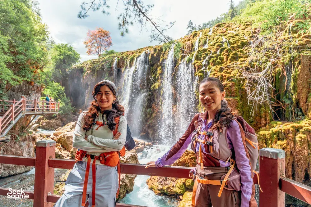Two travellers pose at a wooden railing by a multi-tier waterfall near Shangri-La during a 6-day trek loop