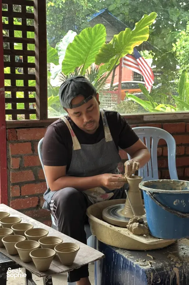 Ceramic artist at Ilham Ceramic in Langkawi works on a pottery wheel, with finished cups on a tray beside a sunlit window.