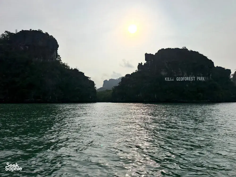 Sunlit waters in Kilim Geoforest Park, Langkawi, with jungle-clad cliffs and a shimmering wake from a cruise boat