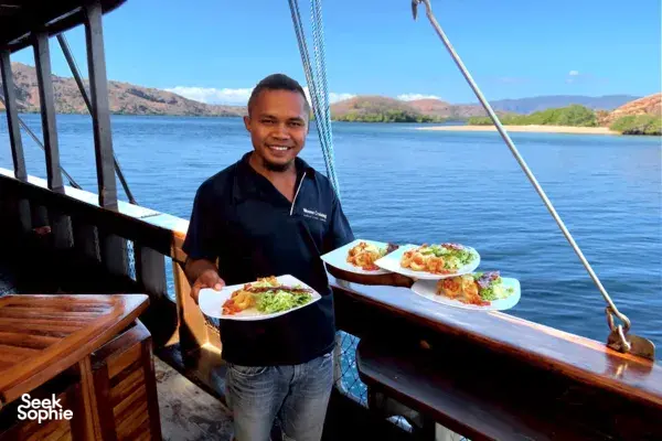 Crew member plates up lunch on the liveaboard deck as calm waves roll by, with mountain views near Labuan Bajo
