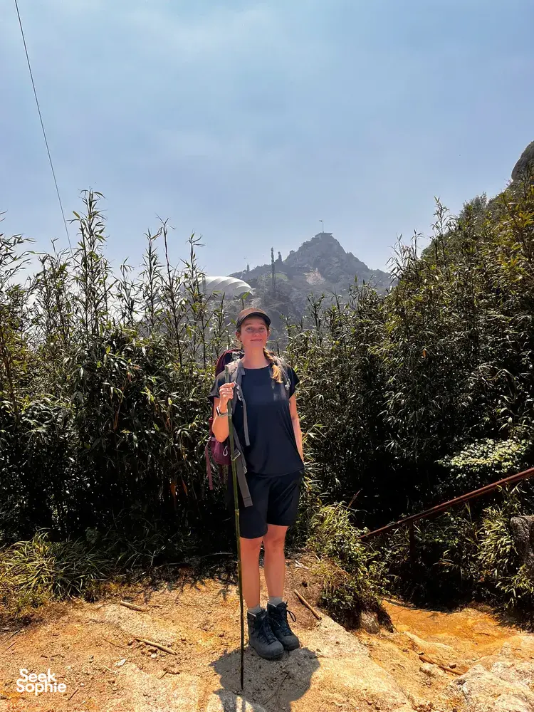 Solo trekker stands on a sunlit path in thick jungle near Mt Fansipan in Sapa, holding trekking poles during the day hike