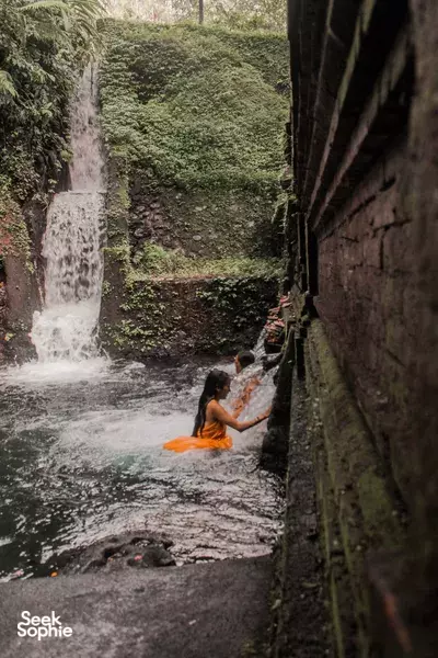 Yoga participant in a narrow waterfall stream in Indonesia, seated on an orange mat with arms raised in a practise pose