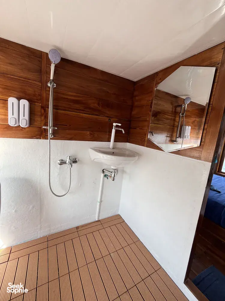 La Famille ensuite on a wooden phinisi showing a countertop sink, overhead shower rail, and mirror as the cruise sails Labuan Bajo
