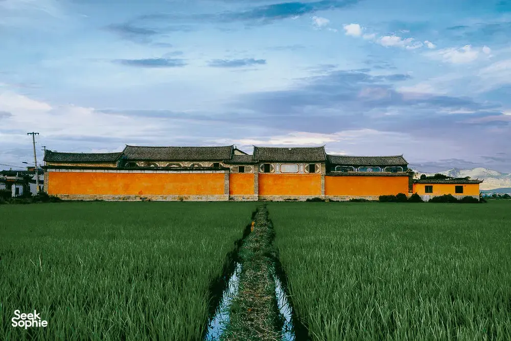 Bangunan warisan gaya Bai berwarna jingga dan hijau di tepi sawah di Xizhou, Dali, dengan banjaran gunung di latar jauh