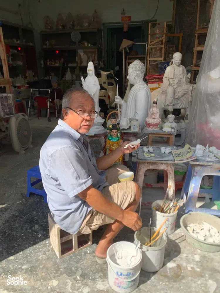 Elderly artisan painting small figurines and surrounded by white Buddha sculptures at a craft shop in Ho Chi Minh City