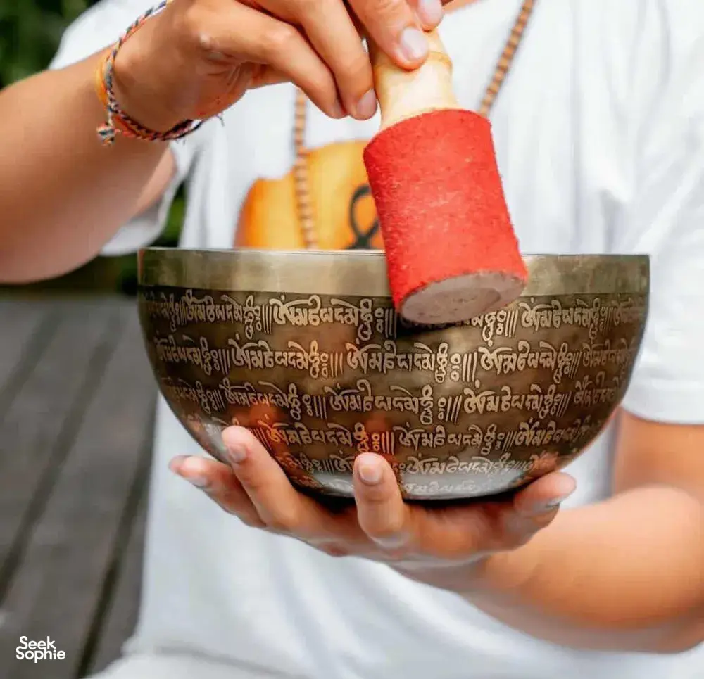 Close view of hands ringing an embossed singing bowl with a red mallet during sound healing on an outdoor mat in Canggu