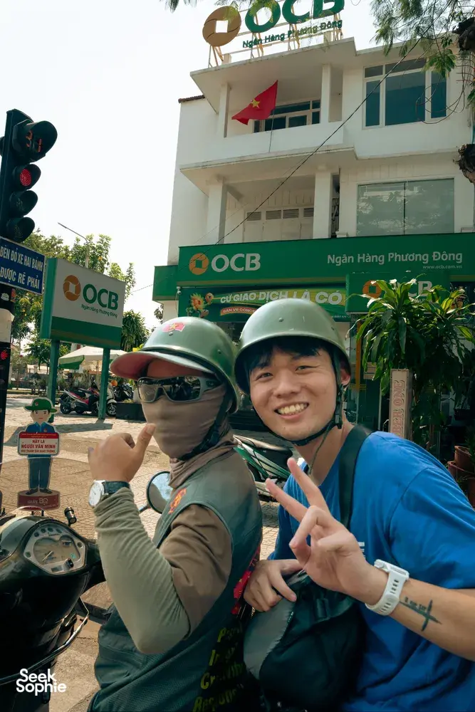 Dua pengendara berhelm di atas skuter vintage di Hoi An, tersenyum dan berpose di samping bendera Vietnam di pusat kota