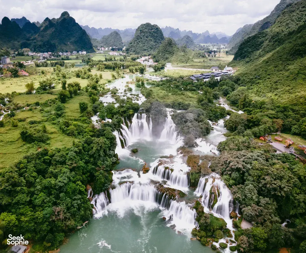 Aerial view of a valley and river with scattered homes, where a broad waterfall cascades through dense green hills