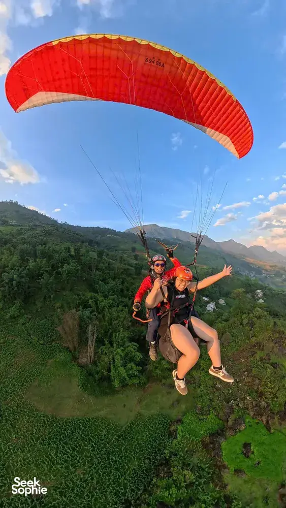 Paragliding passengers float together beneath an orange canopy above forested slopes and distant mountains in Sapa