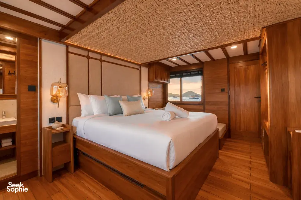 New phinisi bedroom on the deck near Labuan Bajo, with a made-up double bed, wooden walls and sea-view windows