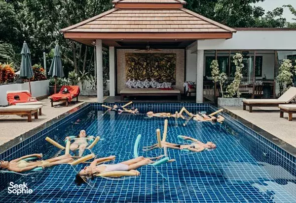 Guests floating and stretching in a tiled swimming pool beside cushioned sunbeds at Phuket Cleanse in Phuket