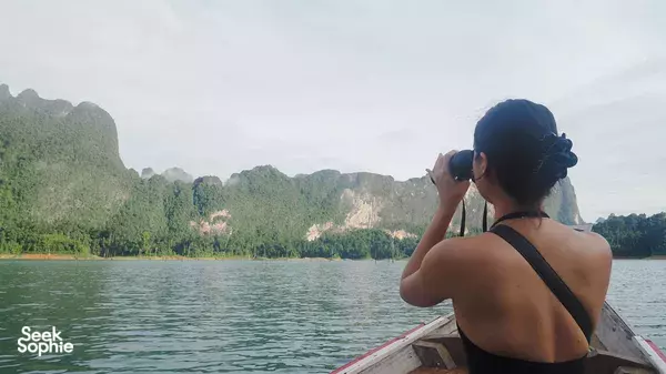 Woman on a boat deck photographing karst peaks across Khao Sok water, with misty mountains behind her on Cheow Lan Lake