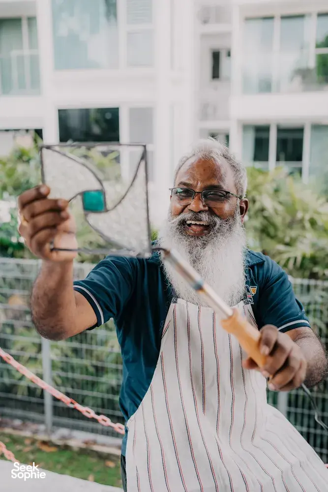 Smiling glass artist in an apron holding a patterned glass panel outdoors in Singapore, centrepiece craft—workshops in Singapore