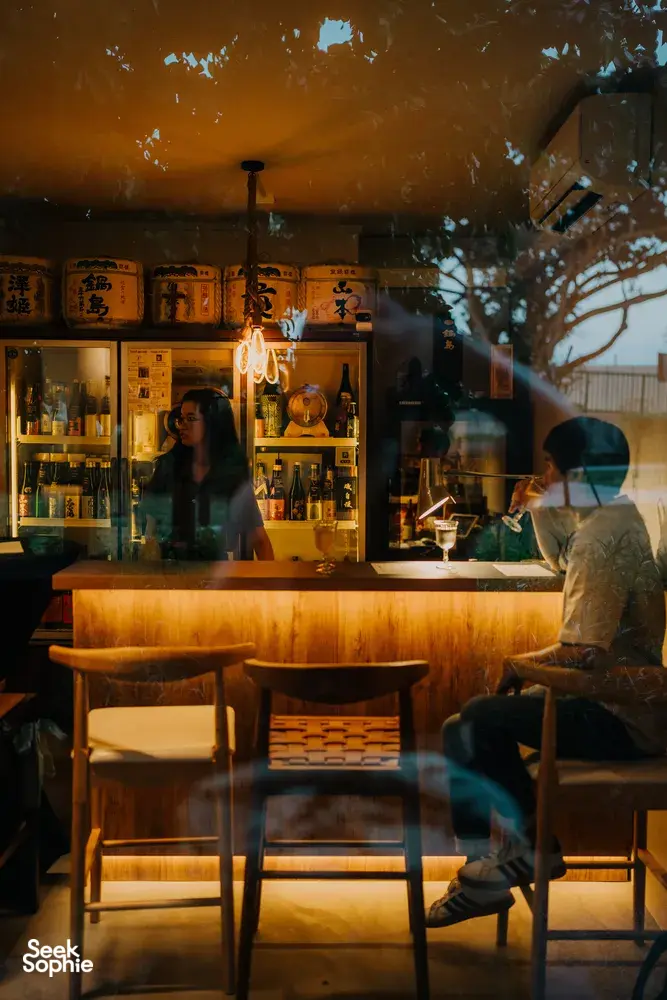 Warm-lit sake bar interior with rows of bottles on wooden shelves and pendant light, in SakeMama’s tucked-away Singapore shop