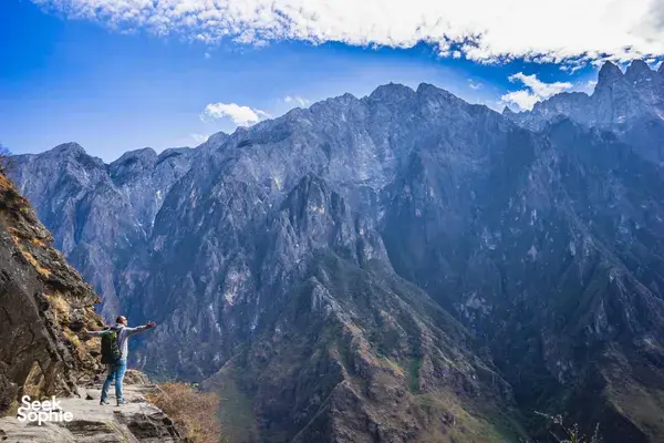 Hiker standing on a rugged cliff path with arms out, looking over steep mountain ranges and valleys in China