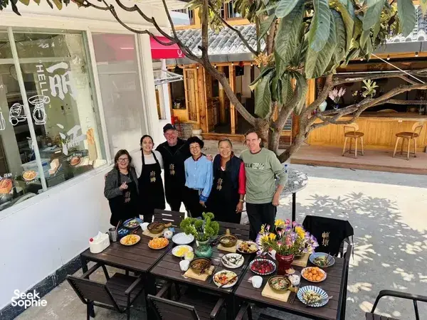 Group of diners eating outdoors in a shaded courtyard, with a spread of Chinese dishes on low wooden tables