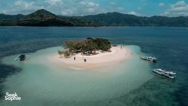 Aerial view of a small sandy island, mangroves and anchored boats in a turquoise bay off Lombok with hills inland