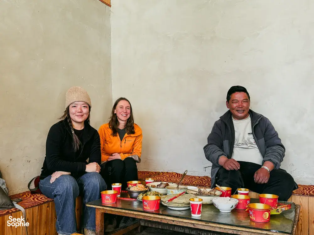 Three guests seated indoors around a shared meal with red cups at a Lijiang guesthouse during a Yi & Naxi village trek