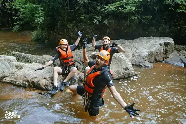 Canyoning in Vietnam: canyoneers in orange life vests pose and wave from a shallow rocky pool surrounded by jungle