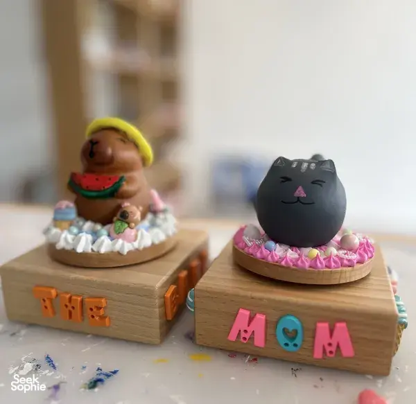 Painted cartoon cat and doll figures on two wooden musical-box bases on a crafting table at Sage Artelier, workshops in Singapore