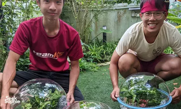 Two friends kneeling in a lush secret farm garden beside large glass terrariums, participating in a terrarium workshop in Singapore