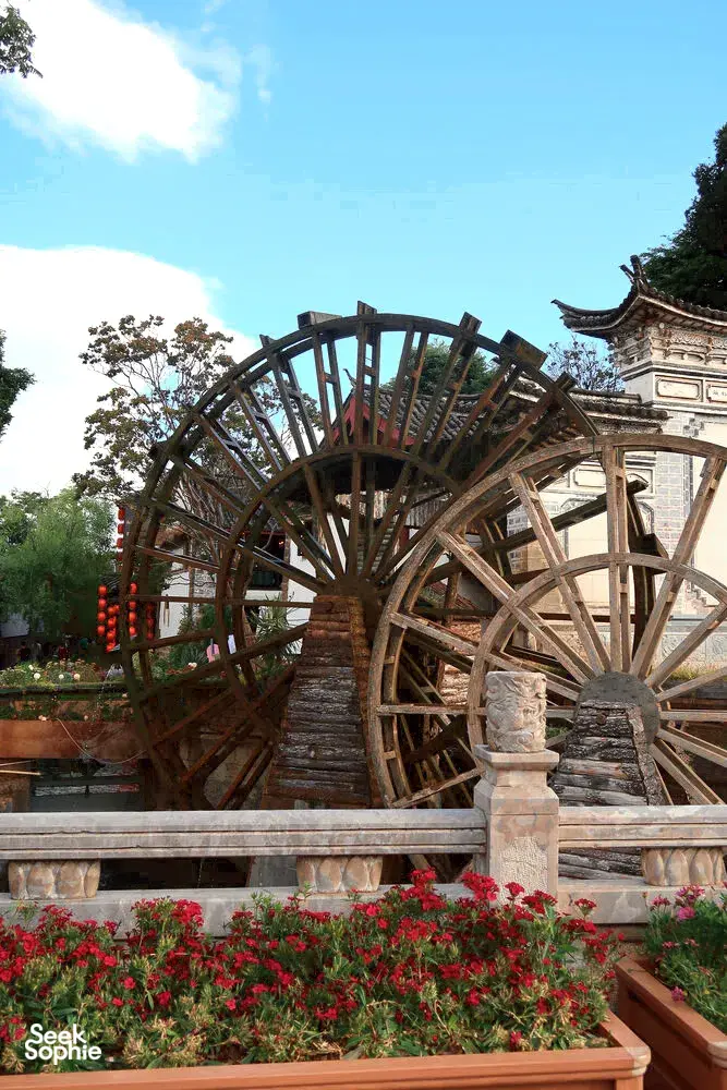 Wooden waterwheel over stone steps with blooming flowers in Shuhe Ancient Town, Lijiang, beside traditional Naxi architecture