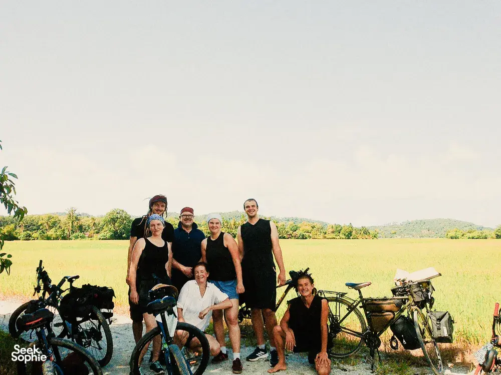 Bicycle Day Langkawi group photo in a sunlit field on Langkawi, posing with bicycles and camping gear by the roadside