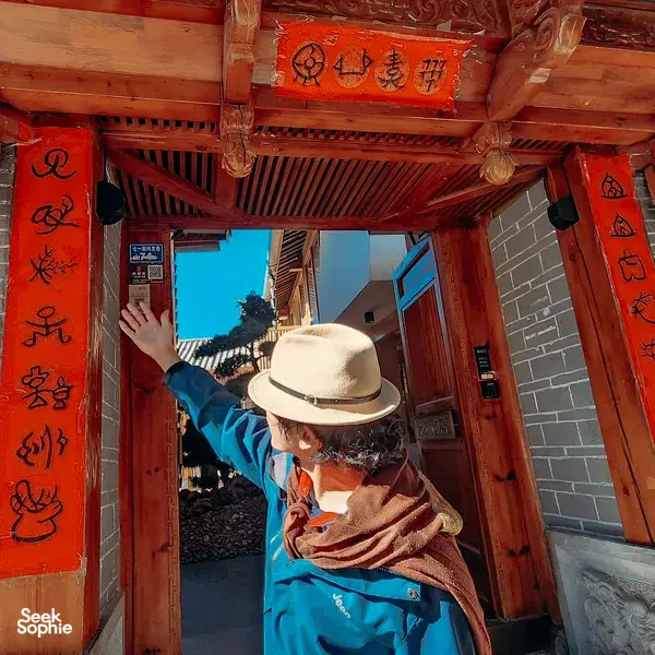 Lijiang Old Town entrance corridor with a traveller in a wide-brim hat pausing beside red-carved wooden doors