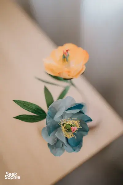 Close-up of two lifelike paper flowers, orange and blue, with green leaves on a beige wall in Singapore studio