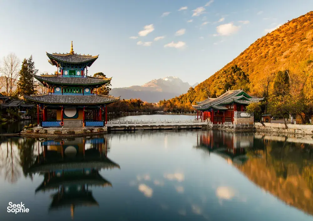 Pavilion dan gerbang bersejarah di tepi tasik merentasi air yang tenang, dengan menara dan bukit musim luruh yang bersinar pada waktu keemasan di Utara Yunnan