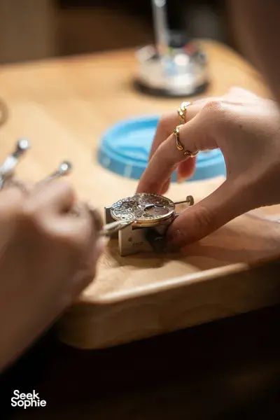 Close-up of hands fine-tuning a watch movement on a workbench while a silver loupe rests nearby — watchmaking workshops in Singapore