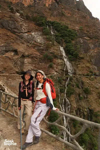 Two hikers with backpacks pause by a barrier overlooking a waterfall in Tiger Leaping Gorge near Lijiang on the 2D1N high trail