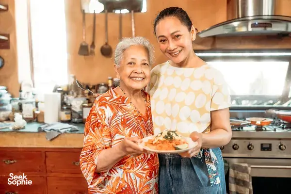 Chiang Mai kitchen cooking class moment: two women smiling while holding a bowl of prepared food in front of the stove.