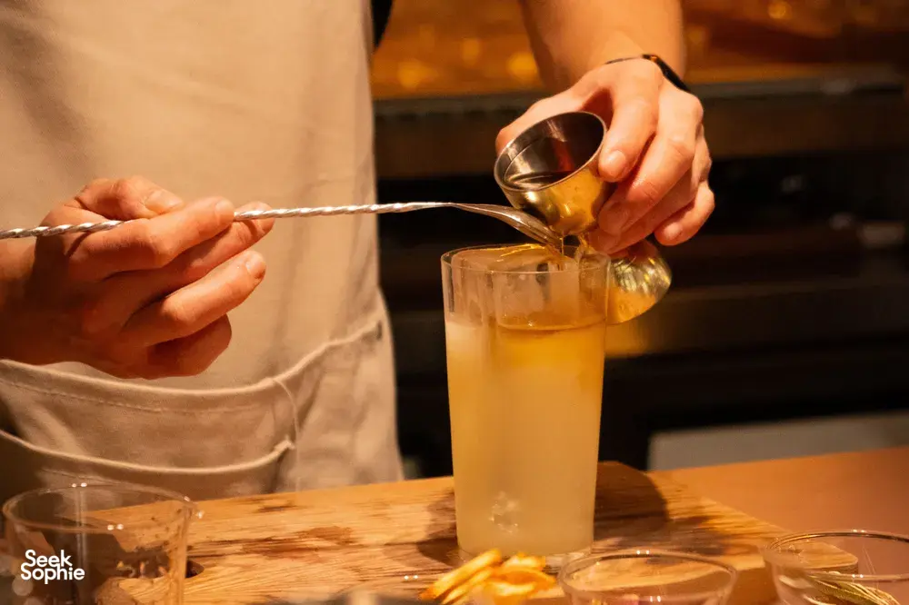 Bartender pours an amber mixer into a tall glass at a Singapore cocktail bar, with a long stir spoon in hand