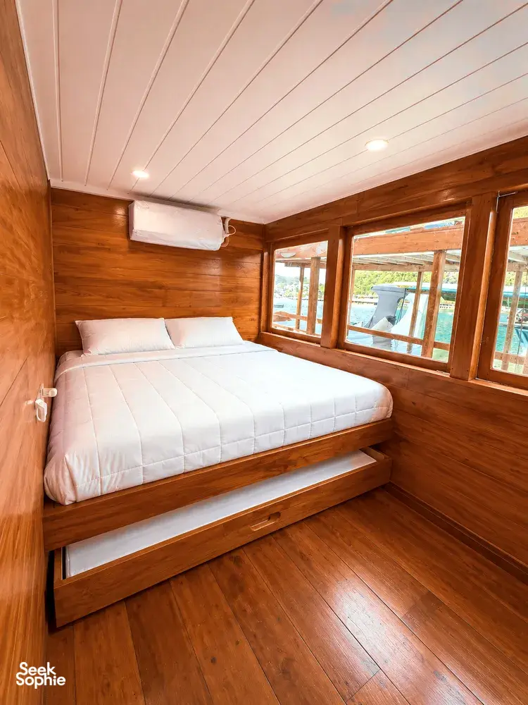 Wooden phinisi cabin with a double berth and side window inside a 3D2N shared Komodo cruise, Komodo Island