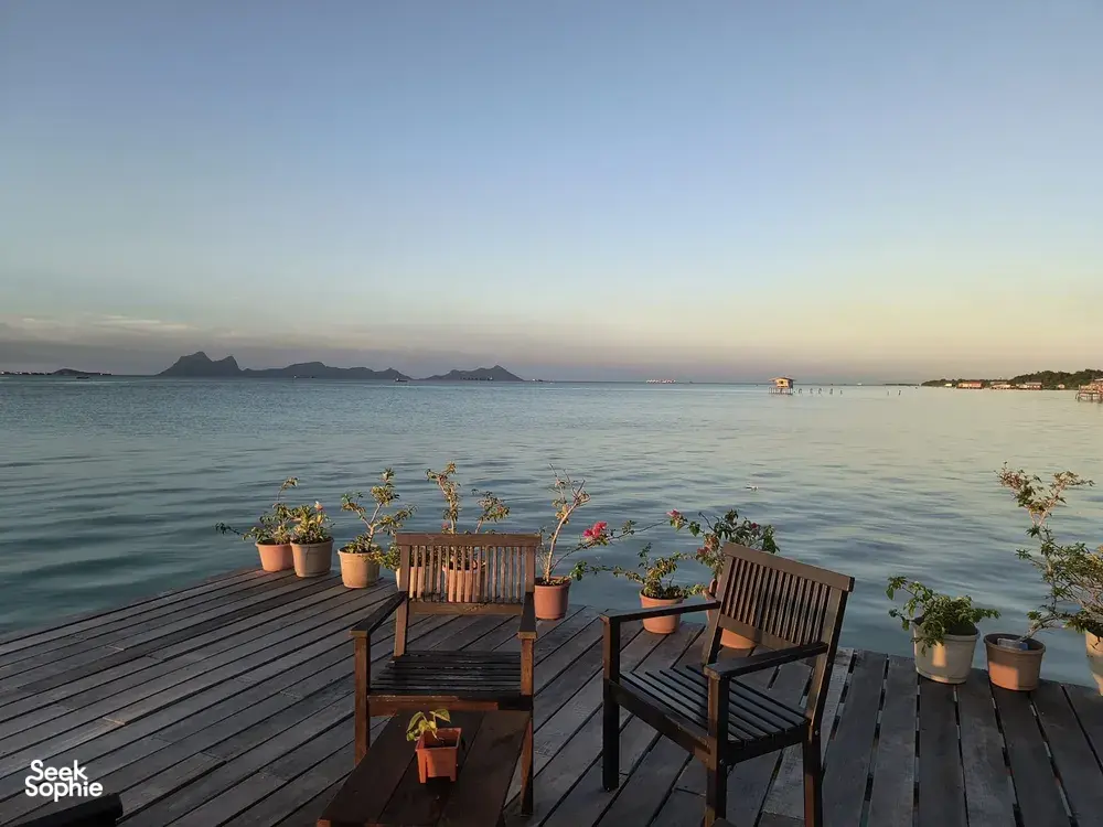 Tempat duduk kayu di dek banglo atas air di Semporna pada waktu senja, dengan tanaman pasu, laut yang tenang, dan pulau-pulau di kejauhan