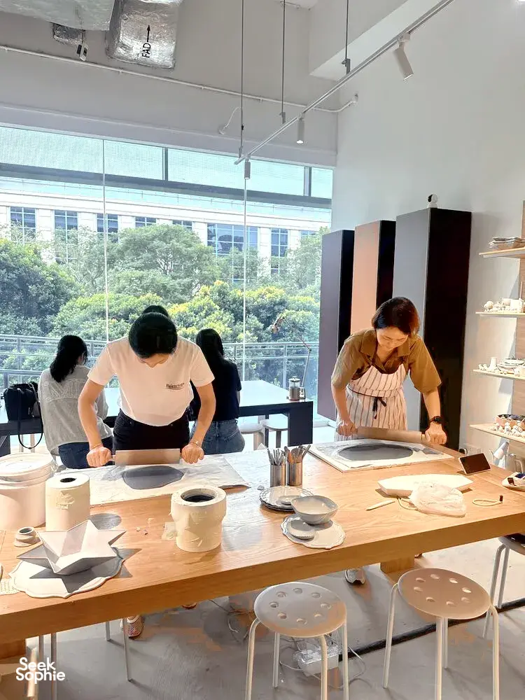 Para peserta di meja kerja tembikar membentuk hasil seramik di sebuah studio cerah di Singapura, dengan peralatan tersusun dan tingkap besar di belakang