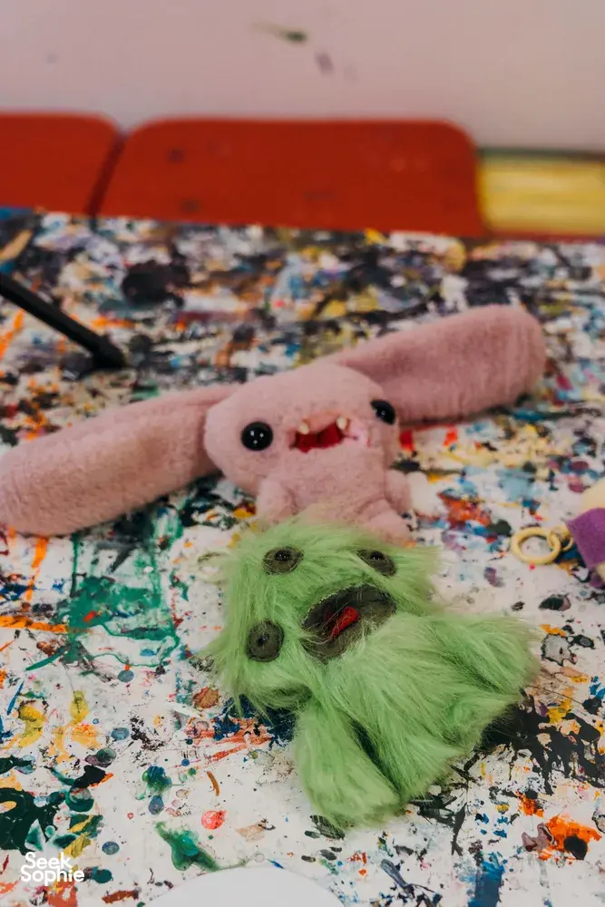 Ugly-cute plushie keychain creations—pink and green plush on a paint-stained table—at Avant-Garde Art Space workshops in Singapore