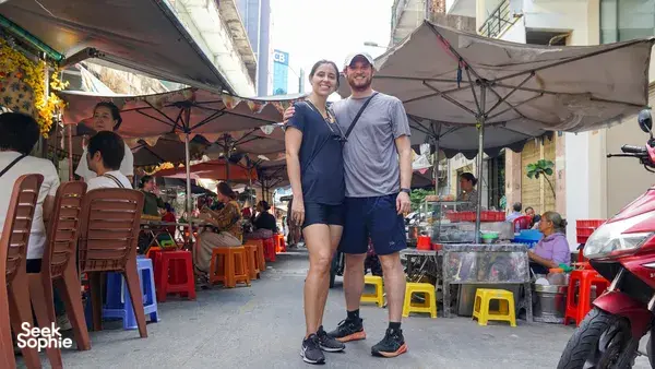 在胡志明市热闹的街市小巷里，两位旅行者撑着伞站着，参加 Jackfruit Adventure 的单车一日行程