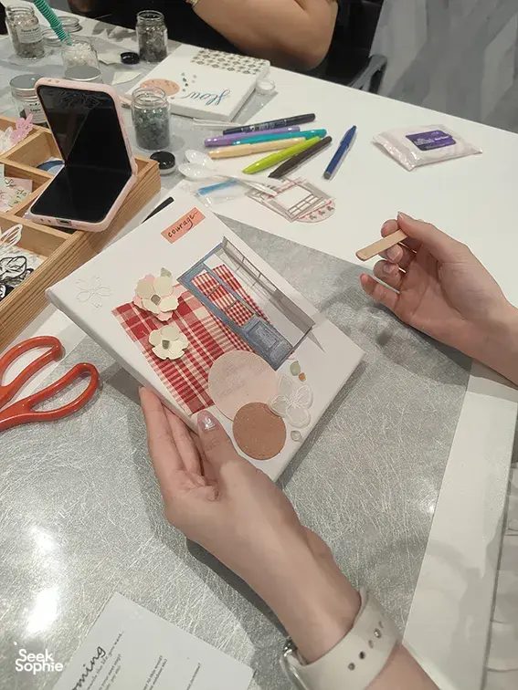 Hands assembling a vision board with gemstone collage pieces at a table in Singapore, with scissors and pens out; workshops in Singapore
