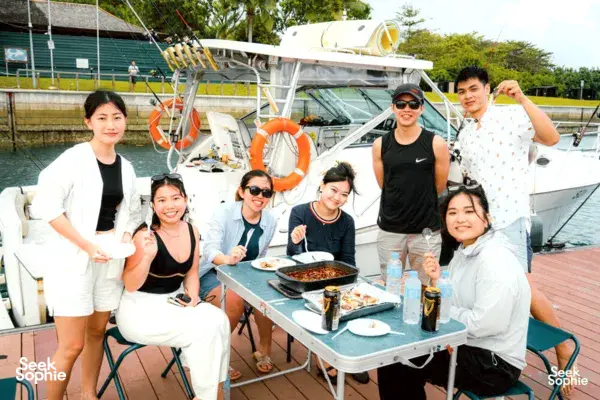 Singapore fishing yacht trip group posing on deck with a tray of food after a catch, with rods and life rings nearby